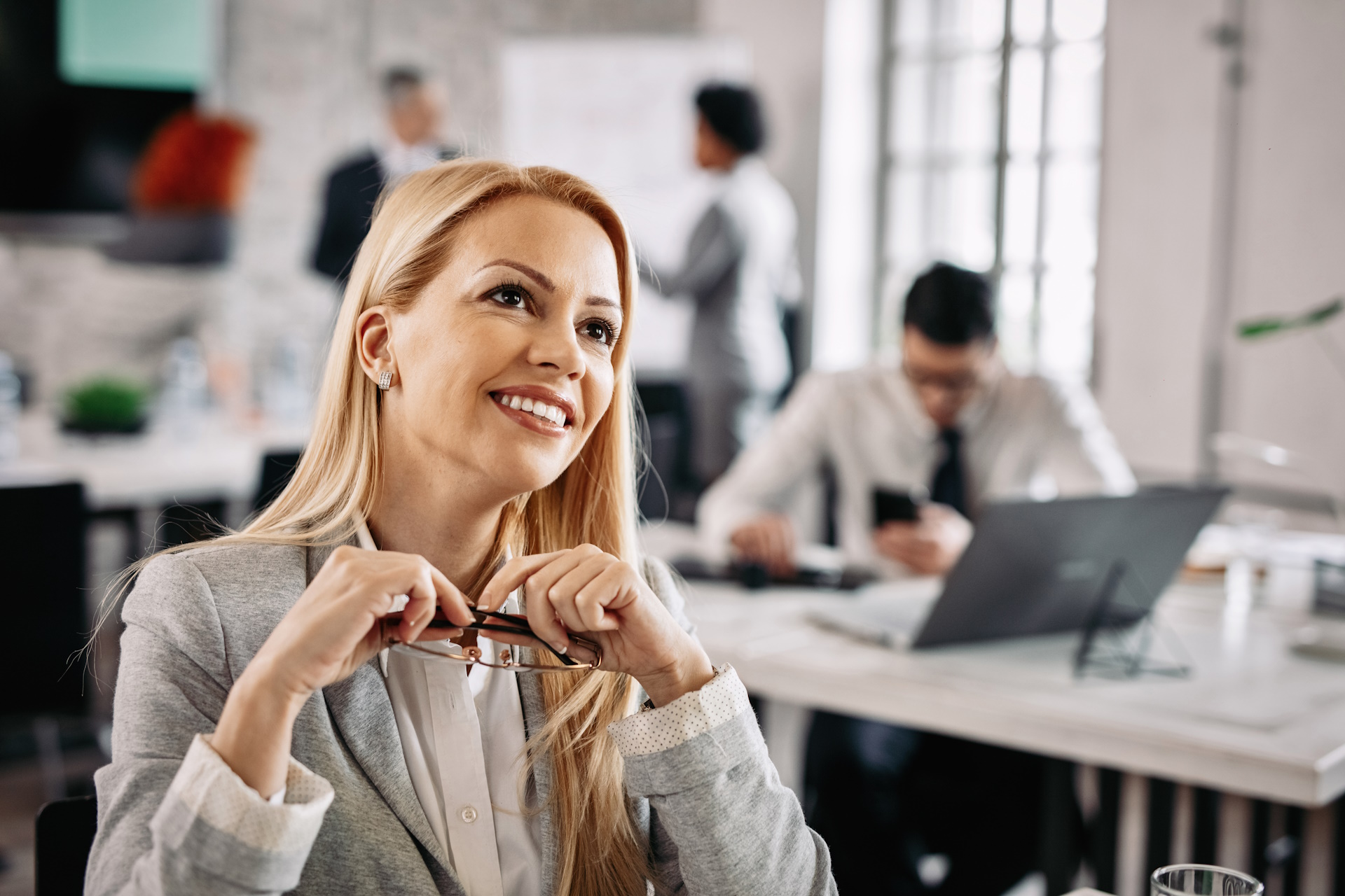 Smiling pensive businesswoman day dreaming while being at work. There are people in the background.